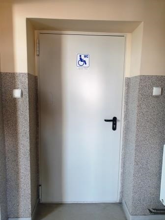 Obraz przedstawia drzwi do toalety dla osób niepełnosprawnych. Drzwi są koloru białego i odpowiednio oznaczone piktogramem.